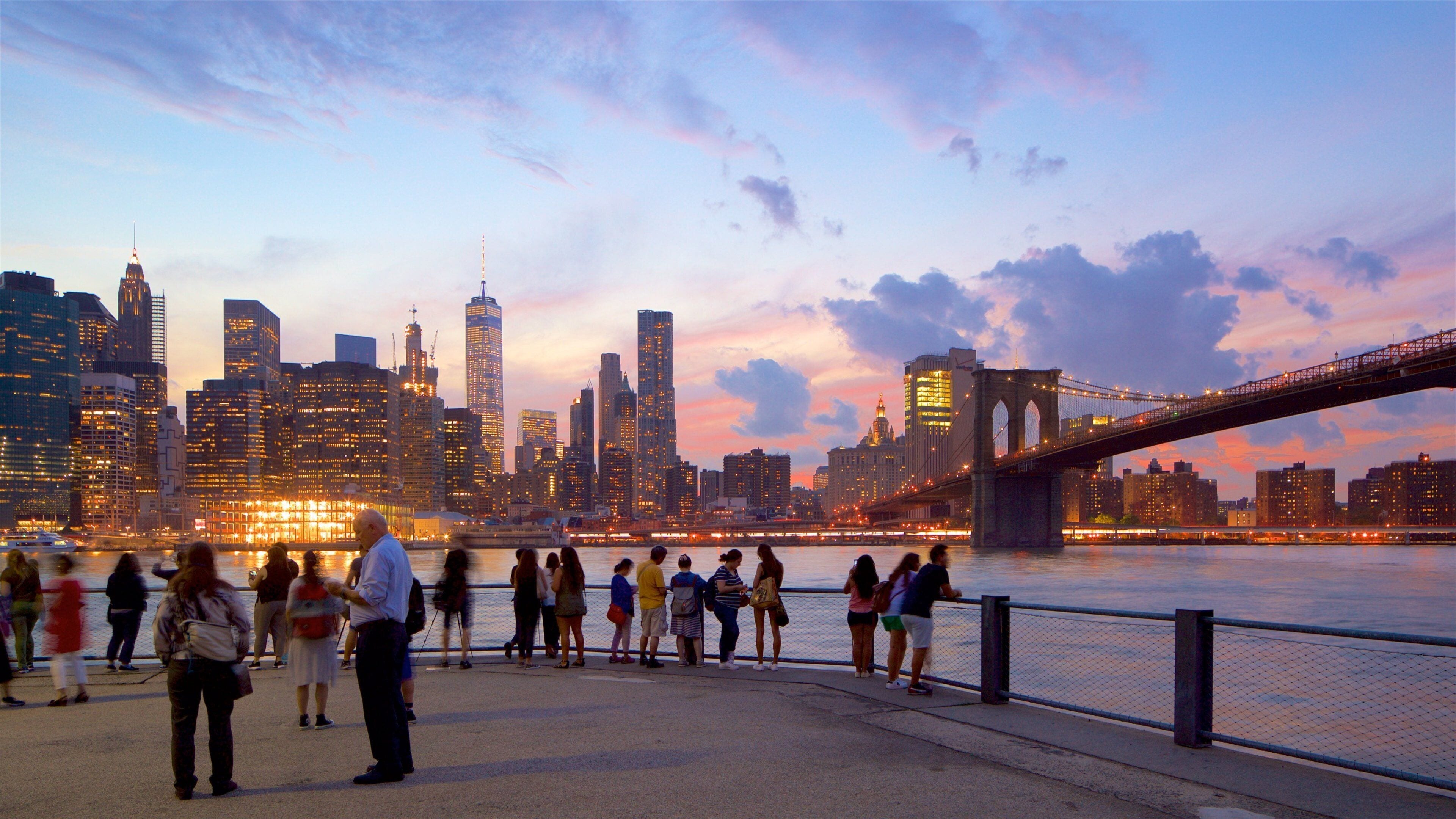 Bezoekers langs de waterkant in Brooklyn met uitzicht op de verlichte Manhattan-skyline en Brooklyn Bridge.