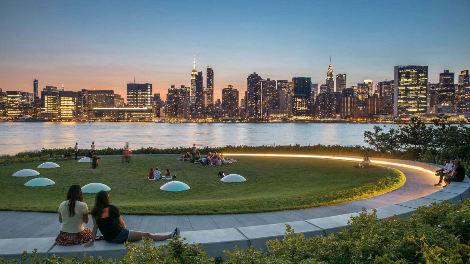 Het ronde grasveld en de zitplaatsen van Hunters Point South Park met uitzicht op Manhattan bij schemering.