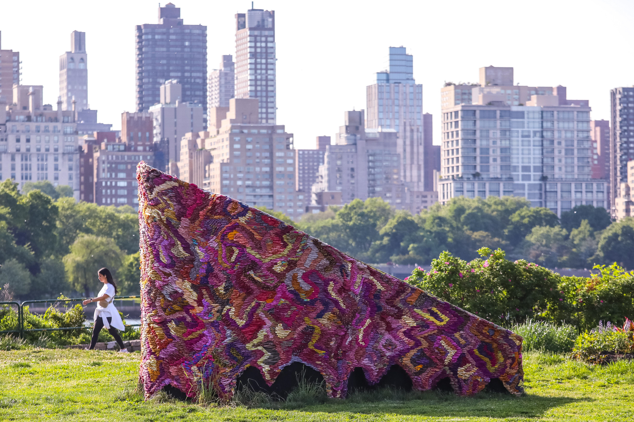 Een kleurrijk driehoekig kunstwerk op het gras van Socrates Sculpture Park, met Manhattan op de achtergrond.