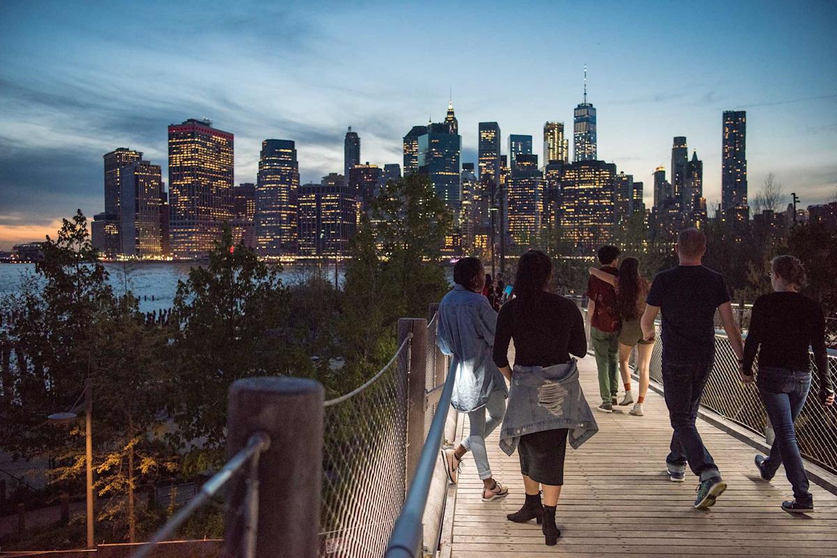 Wandelaars op Squibb Park Bridge met de verlichte skyline van Manhattan bij schemering.