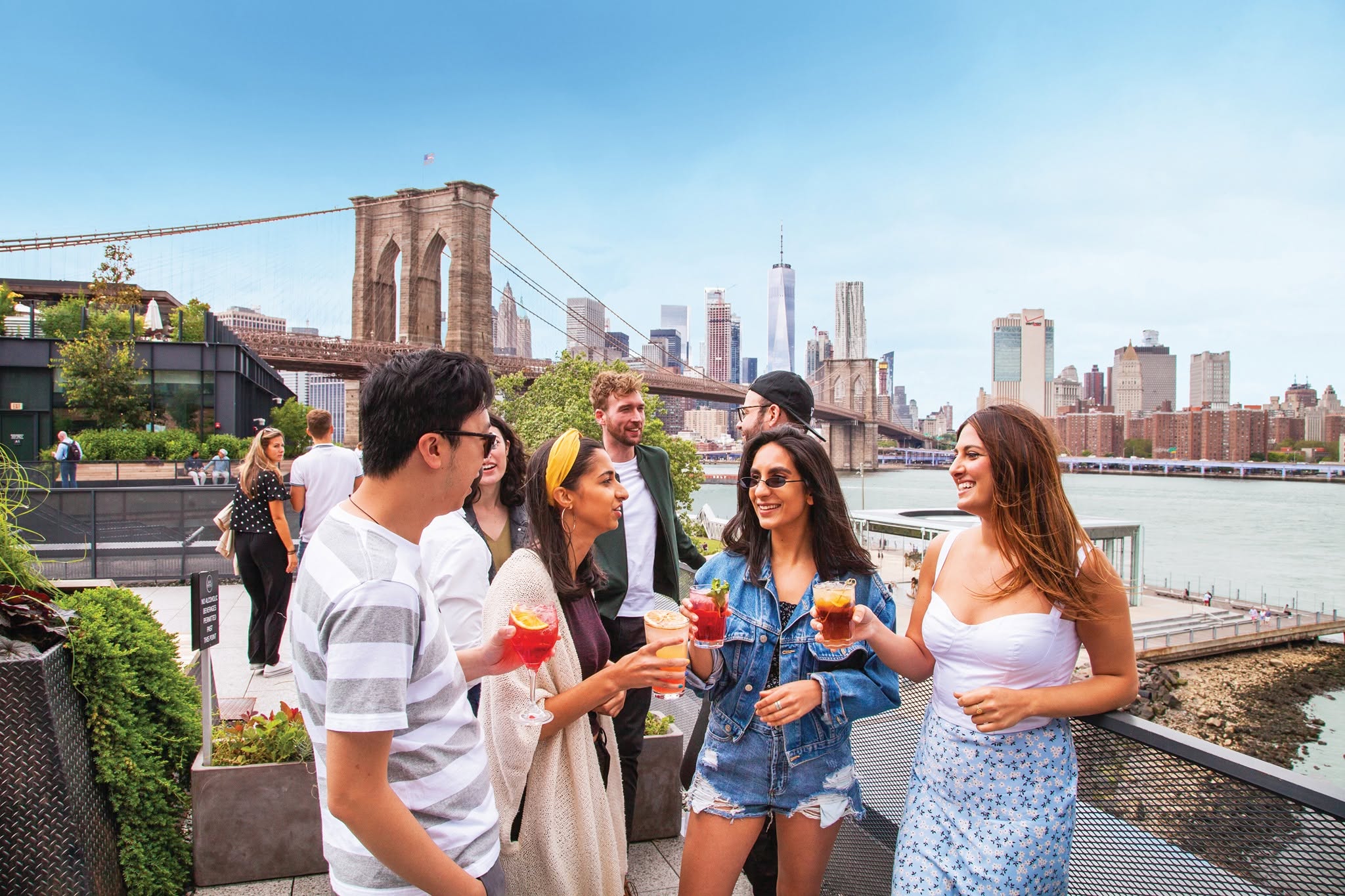 Bezoekers met drankjes op het Time Out Market-dakterras, met de Brooklyn Bridge op de achtergrond.